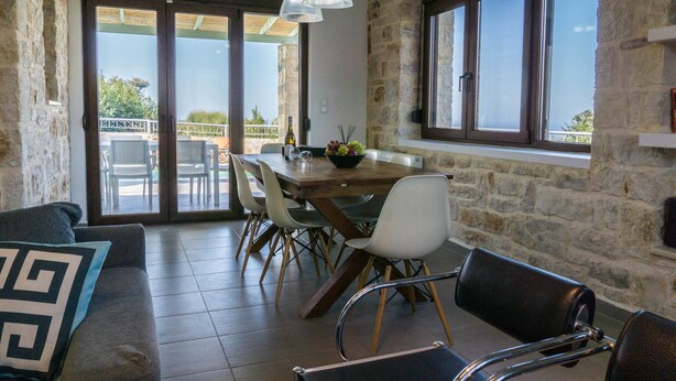 A dining area with a stone wall, a wooden table, white chairs, and a view through large glass doors.