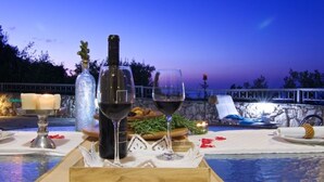 A table set with wine glasses, a bottle, candles, and a tray of food, overlooking a pool area at dusk. - Holiday Home in Crete With Private Pool (Rethymno)