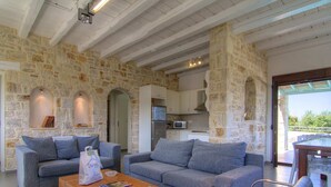 A modern living room with stone walls, a sofa set, a coffee table, and a dining area with chairs. - Holiday Home in Crete With Private Pool (Rethymno)