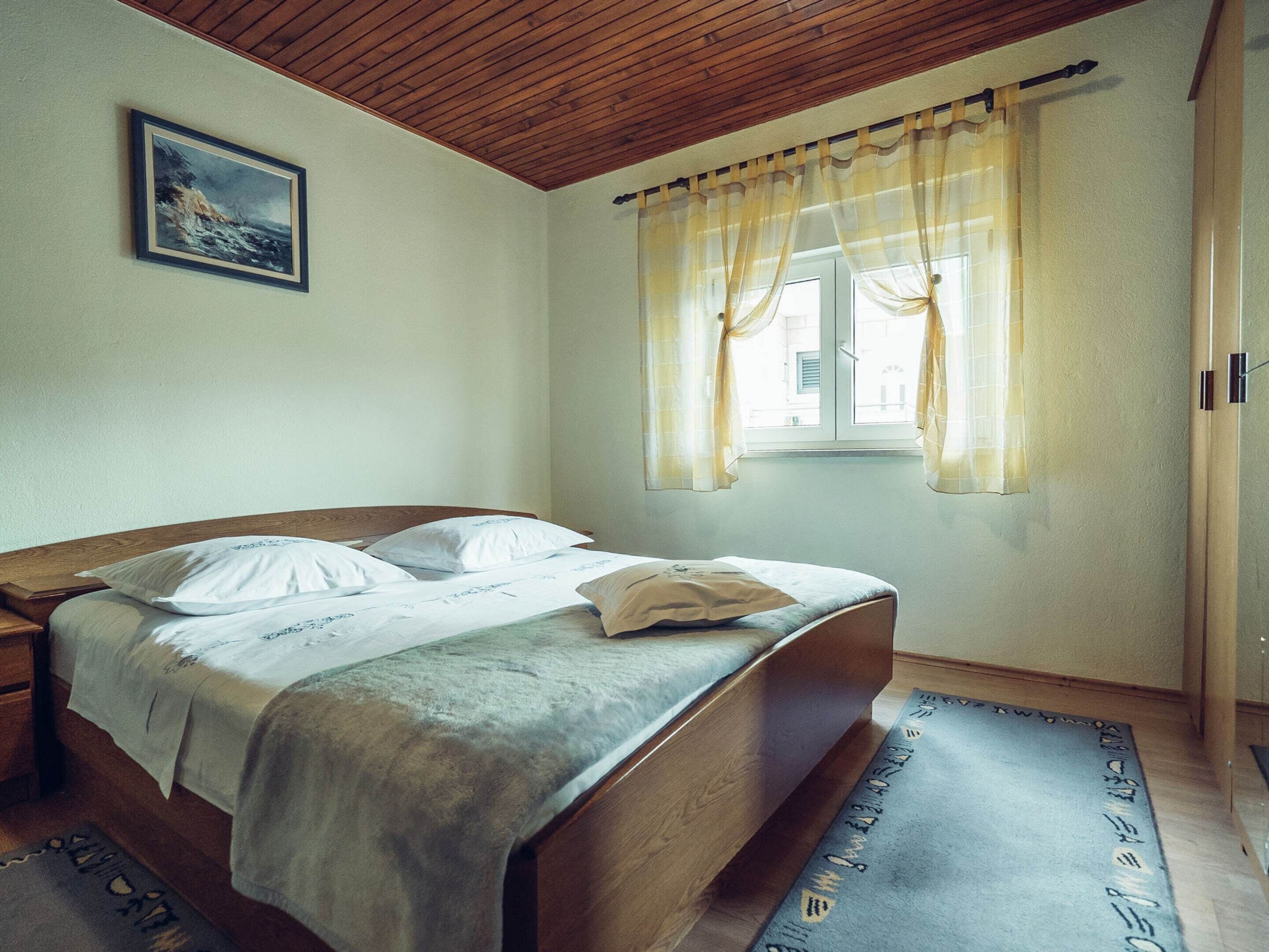Een slaapkamer met een houten bed, een nachtkastje, een raam met gordijnen en een schilderij aan de muur.