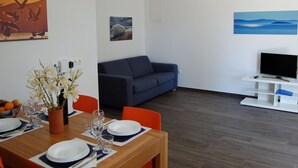 Un soggiorno moderno con un divano blu, un'area pranzo con un tavolo in legno apparecchiato per quattro persone e una TV su un mobile bianco.