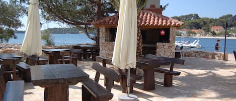 Outdoor seating with tables and benches, a stone building with a tiled roof, and a view of the sea.