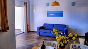 A modern living room with a blue sofa, a dining table with chairs, a vase with yellow flowers, and a painting on the wall.