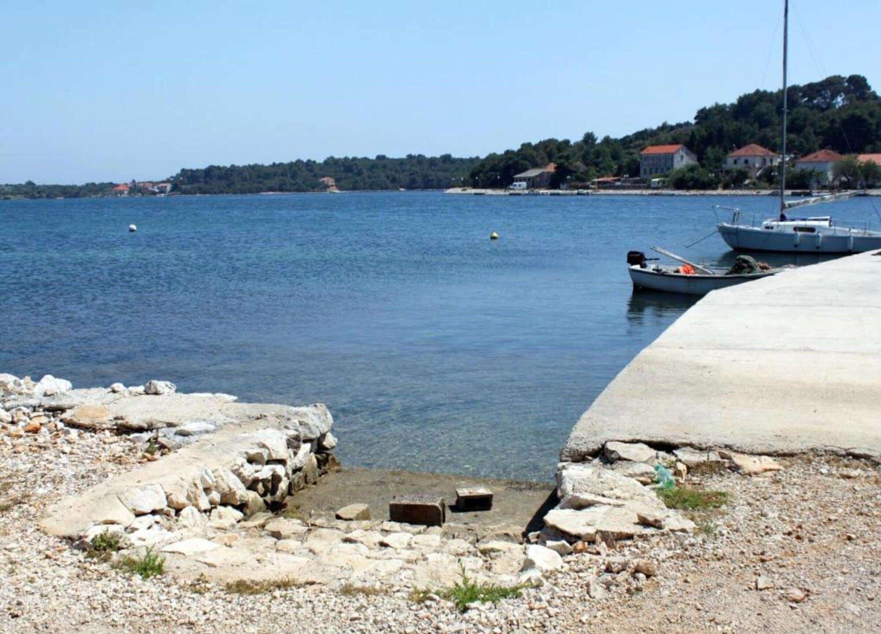 Une côte rocheuse bordée de bateaux amarrés à un quai en béton, une mer bleue et limpide, et une colline lointaine parsemée de bâtiments.