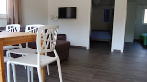 A room with a wooden dining table and white chairs, a brown sofa, a flat-screen TV mounted on the wall, and a bed with a blue mattress in the adjacent room. - Modern Holiday Home With Terrace, Near the Sea (Sali)
