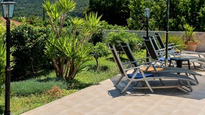 An outdoor seating area with lounge chairs and a large umbrella, surrounded by lush greenery and a view of hills. - Modern Villa in Grohote With Pool (Solta)