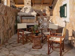 An outdoor seating area with stone flooring, wooden chairs, a wicker table with fruit, and a stone wall. - Modern Villa in Grohote With Pool (Solta)