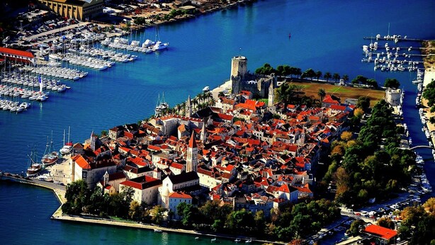 An aerial view of a historic town with a dense cluster of red-roofed buildings, a prominent castle, and a marina filled with boats.