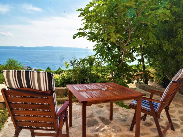 A wooden table and two chairs with striped cushions, set on a stone patio overlooking a scenic view of the sea and distant mountains.
