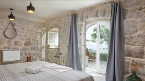 A bedroom with a stone wall, a bed with striped bedding, a mirror, a sink, and a view through a glass door to an outdoor seating area. - Villa in Podgora With Pool Near Beach (Podgora)