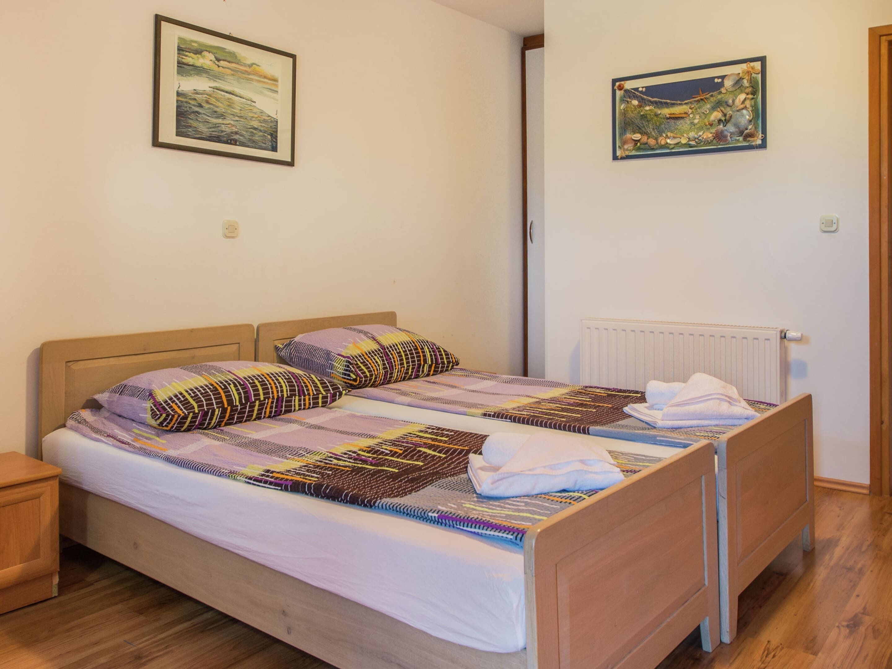 Ein Einzelbett mit Holzbettgestell, zwei Kissen und einer gemusterten Decke. Darüber an der Wand hängen zwei gerahmte Bilder.