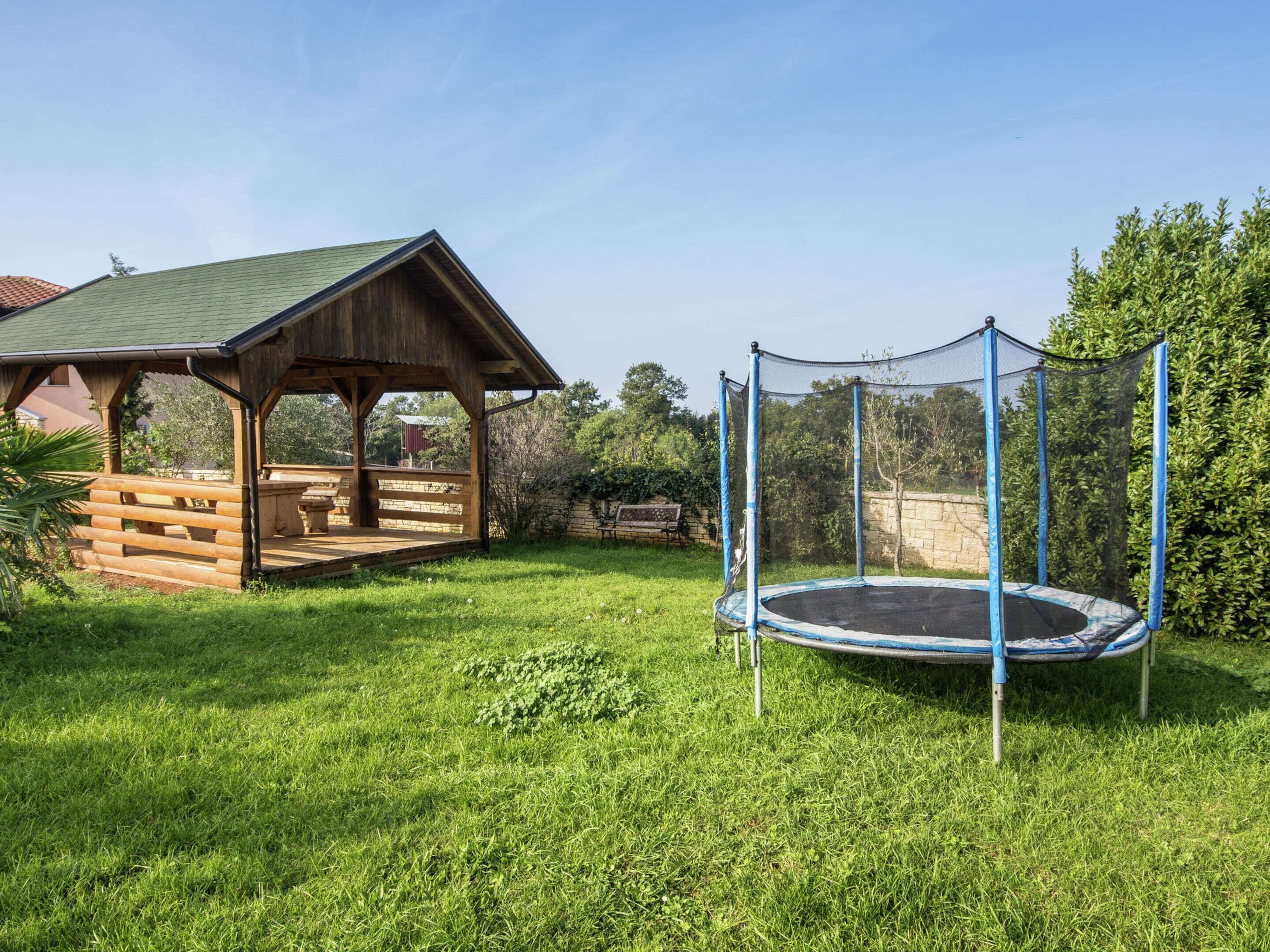 A wooden gazebo with a bench, a trampoline with a safety net, and a grassy yard.