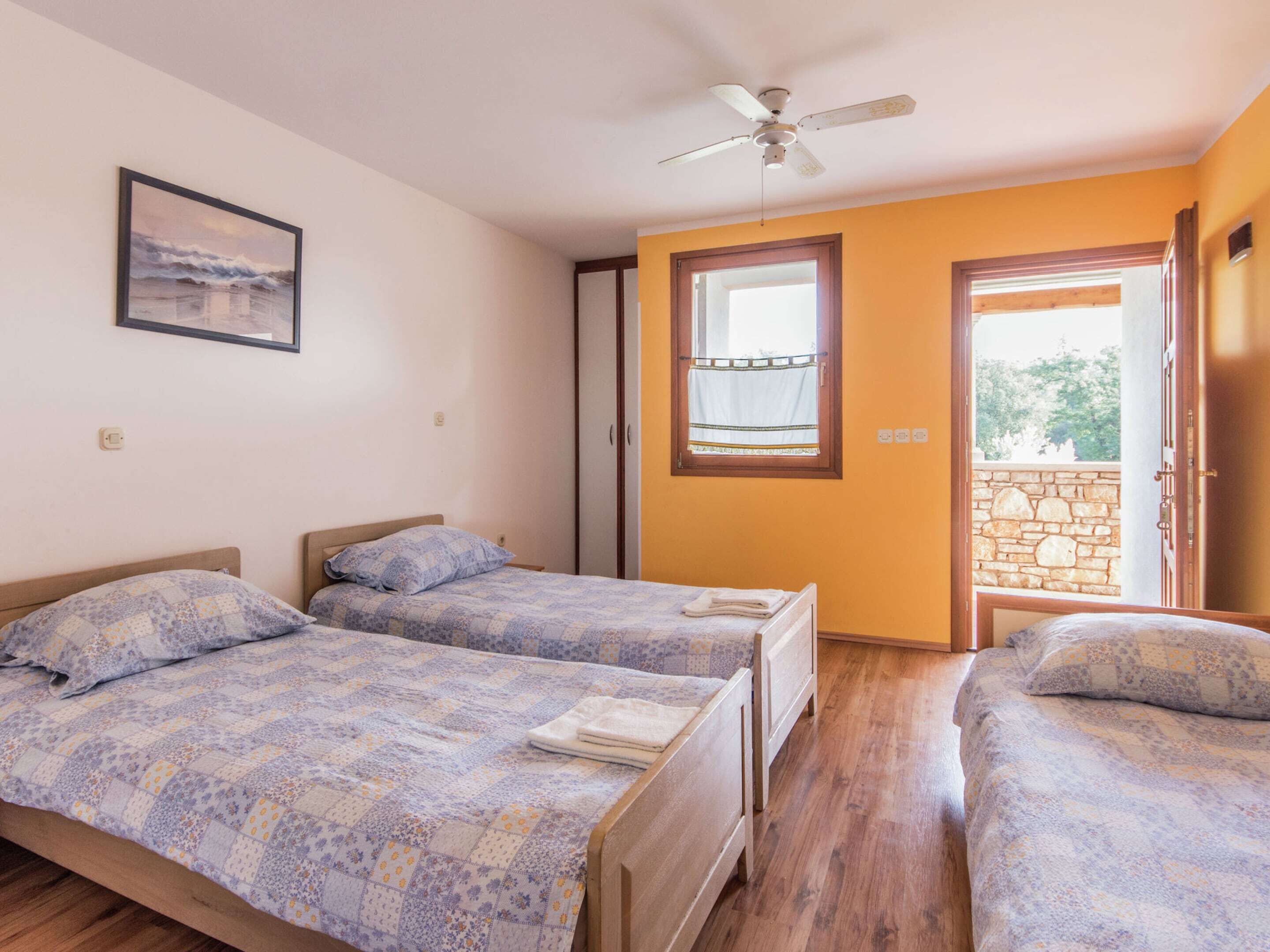Une chambre avec deux lits, un ventilateur de plafond, une fenêtre avec un rideau et une vue sur un mur de pierre à l’extérieur.