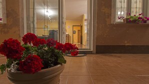 Ein Blumentopf mit einer roten Blume im Vordergrund, eine Glastür, die zu einem Innenbereich führt, und ein Fenster mit Blumenkasten auf der rechten Seite.