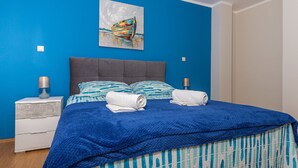Ein Schlafzimmer mit blauer Wand, grauem Kopfteil, einem Bett mit blau-weiß gestreifter Bettwäsche, zwei Kissen und zwei zusammengerollten Handtüchern. An der Wand hängt ein Bild von einem Boot.