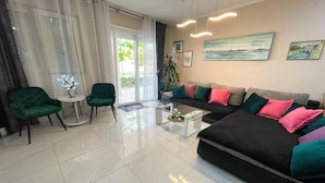 Una sala de estar moderna con un sofá negro, sillones verdes, una mesa de centro de vidrio y vistas al exterior a través de una puerta de cristal.