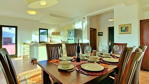 A dining area with a wooden table set for six, surrounded by chairs. A kitchen is visible in the background with modern appliances.