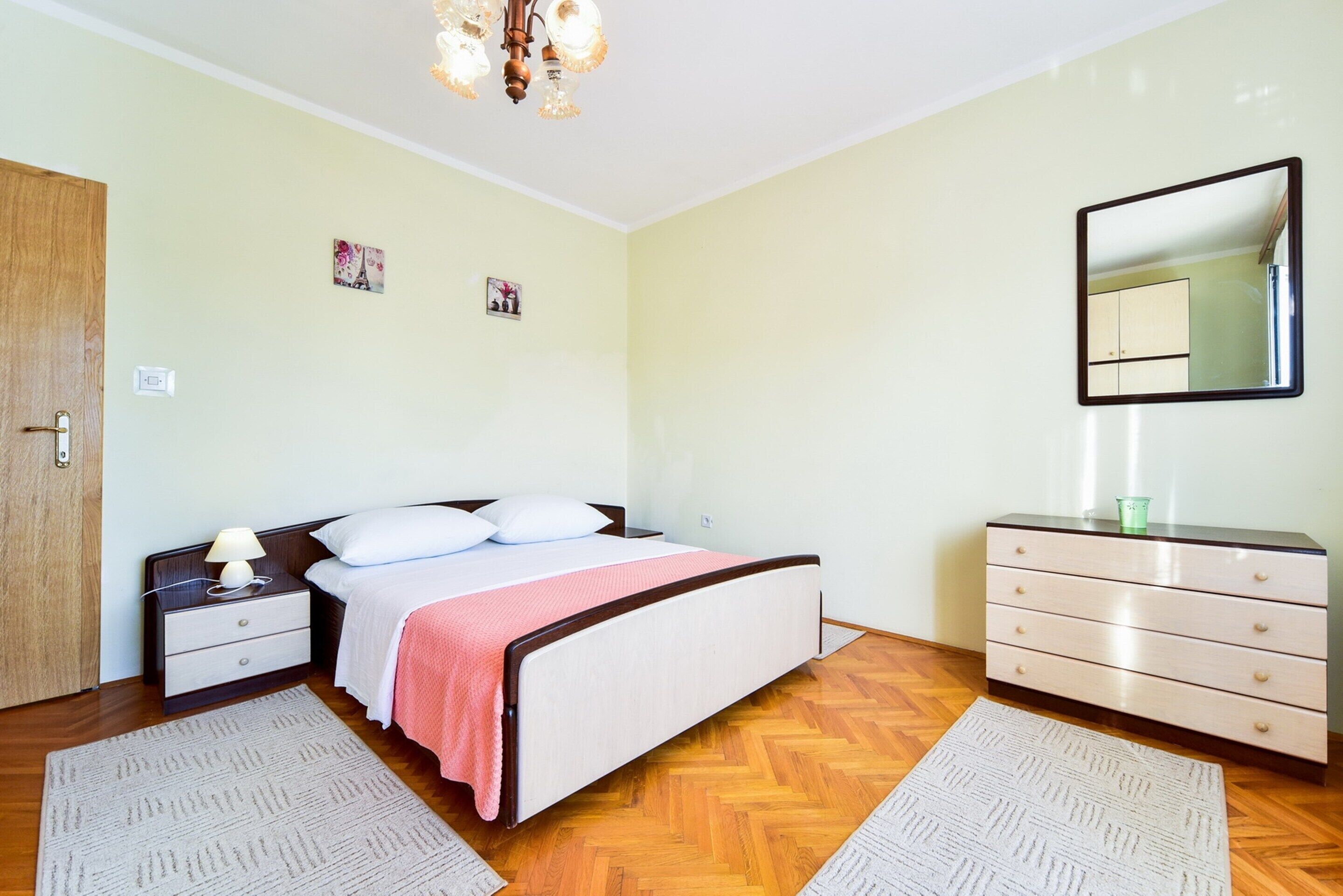 Ein Schlafzimmer mit Bett, Nachttisch, Kommode, Spiegel und zwei Teppichen.