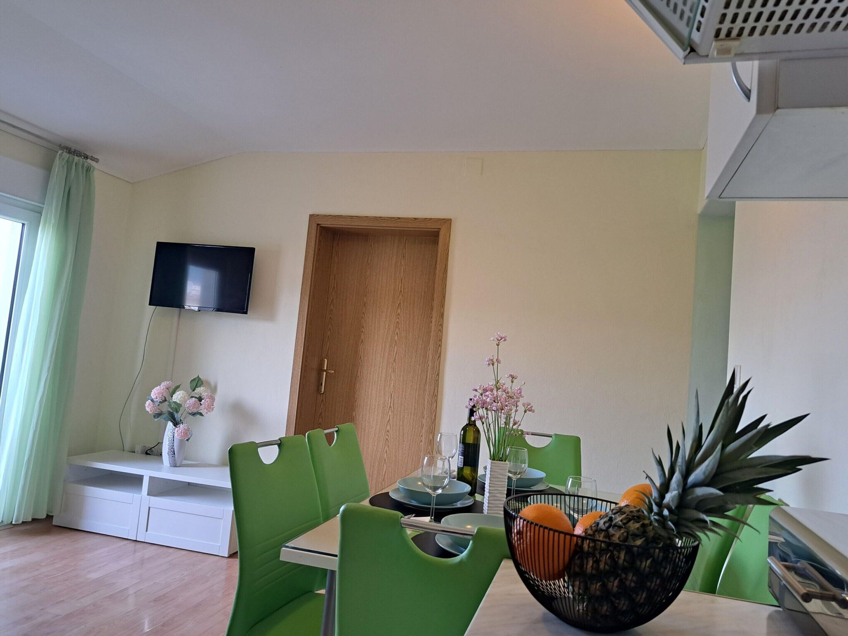 A dining area with a table set for two, green chairs, a flat-screen TV mounted on the wall, a wooden door, and a window with green curtains.