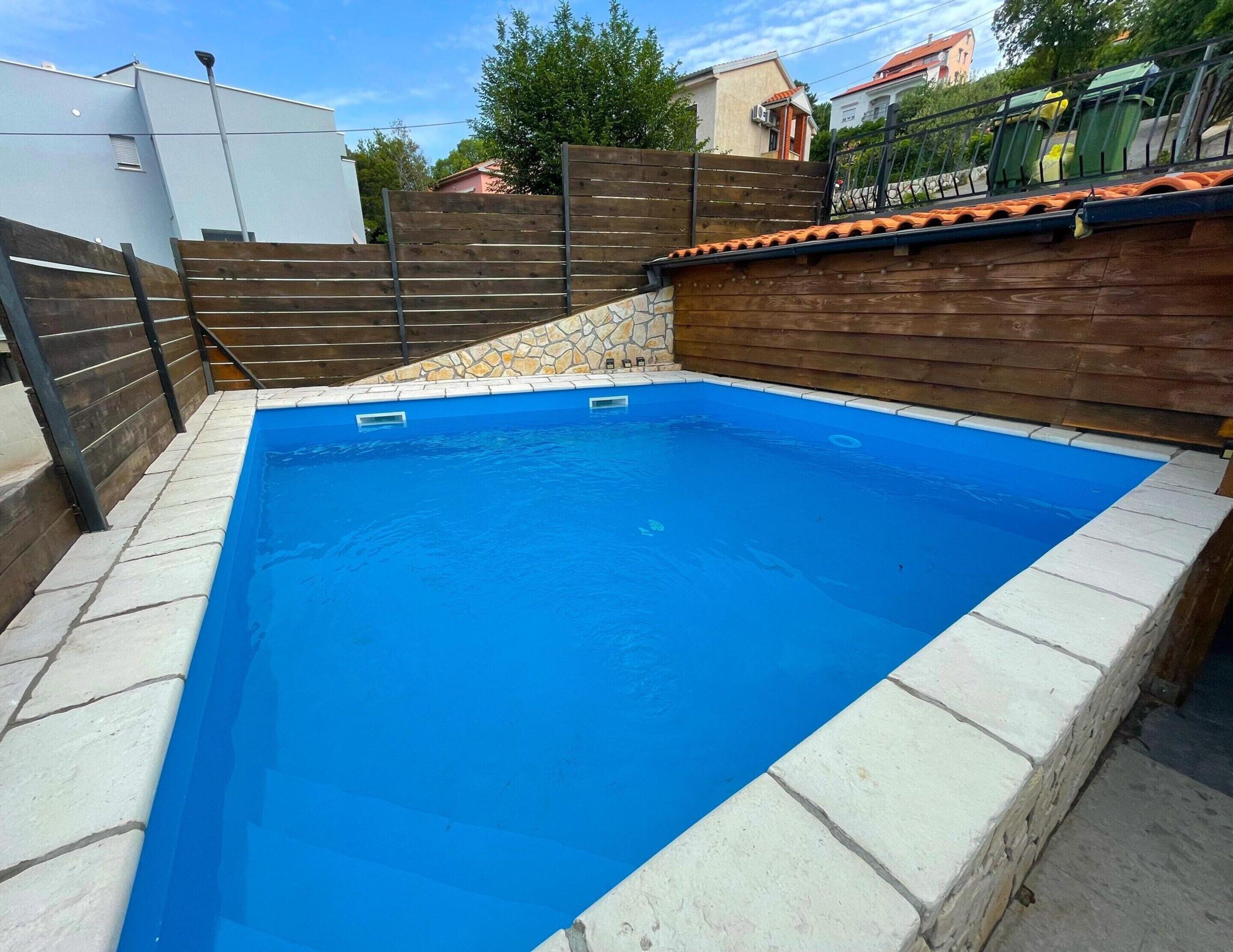 Une piscine rectangulaire bordée de pierres et de carreaux, entourée d’une clôture en bois et d’un mur de pierre.