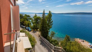 A balcony with a view of a coastal road, trees, and a beach with people. - Studio Apartment With Swimming Pool & Sea View (Crikvenica)