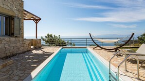 A rectangular swimming pool with clear blue water, surrounded by stone tiles. There is a hammock and a lounge chair on the right side, and a stone building with a window on the left. - Studio Apartment With Swimming Pool & Sea View (Crikvenica)
