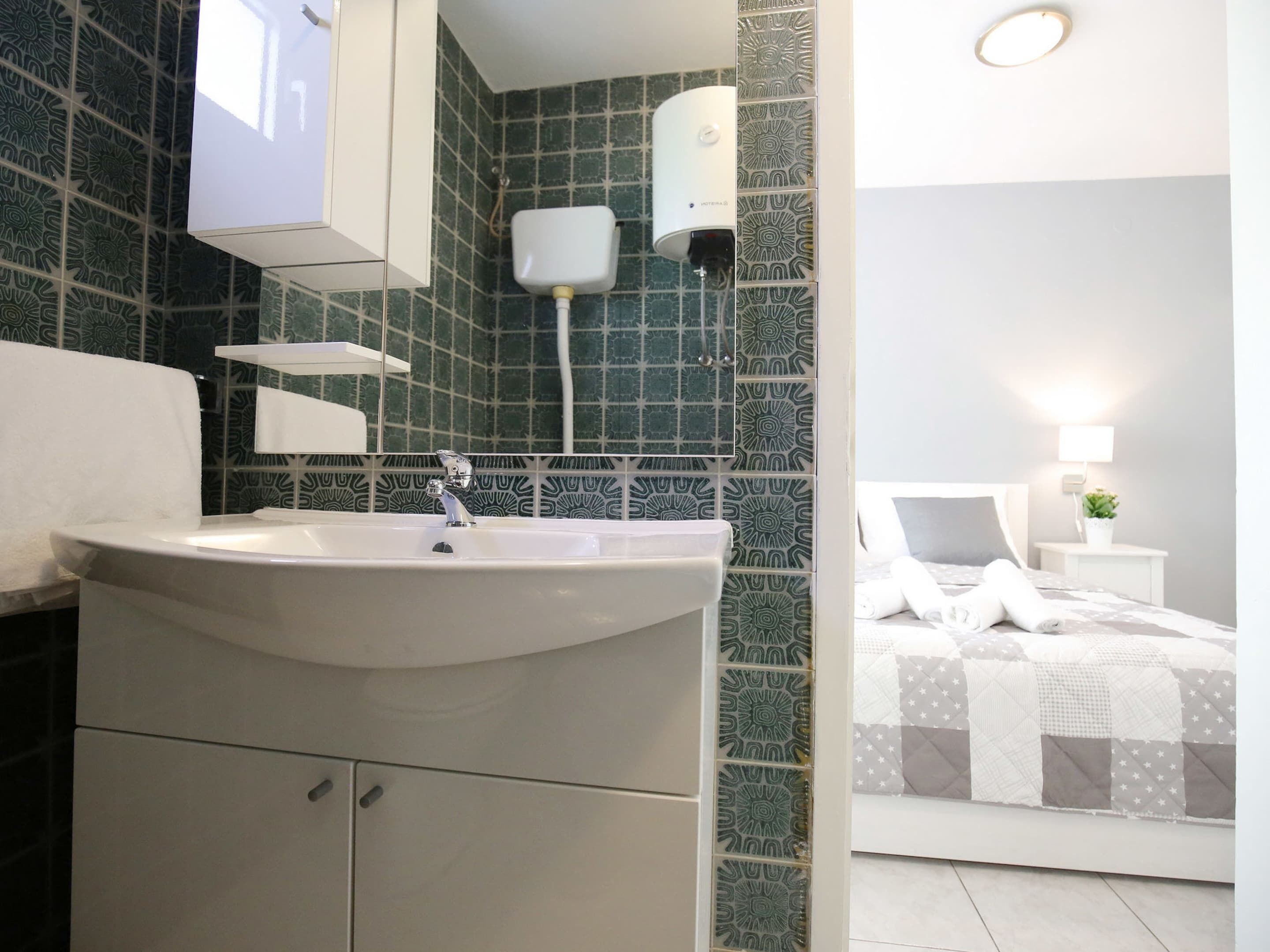 Un baño con lavamanos, espejo e inodoro. En la habitación contigua, hay una cama con mesita de noche y lámpara.
