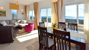 Ein modernes Wohnzimmer mit einer Couch, einem pinken Sessel, einem Esstisch mit Stühlen und großen Fenstern mit Meerblick.