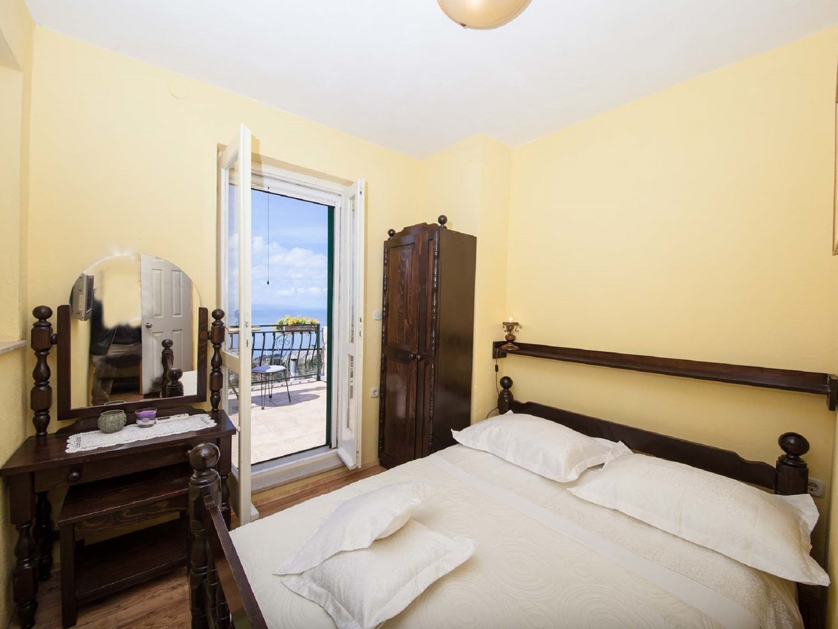A bedroom with a bed, a wooden wardrobe, a dresser, a mirror, and a door leading to a balcony.