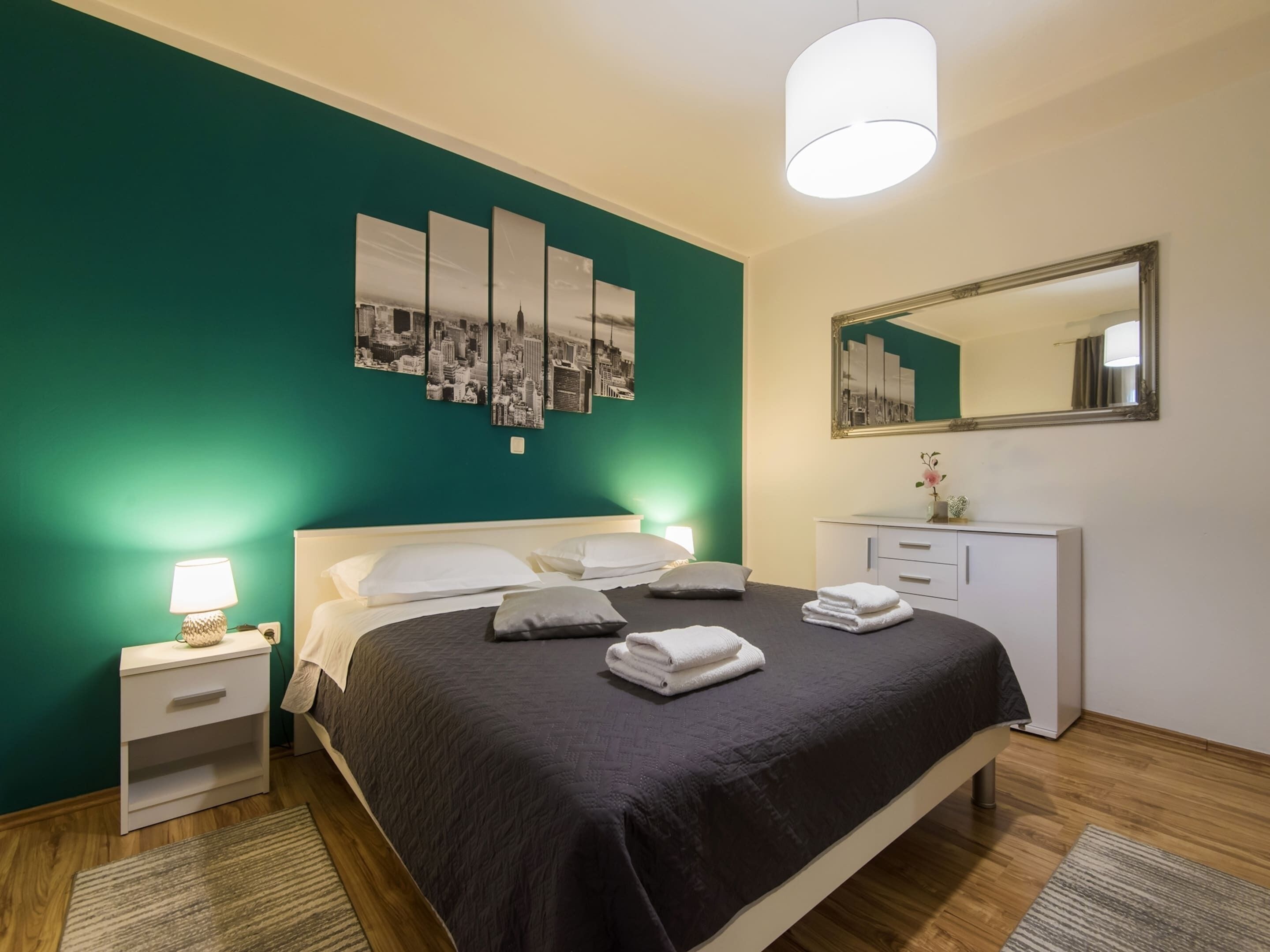Ett modernt hotellrum med en säng, sängbord, en byrå, en spegel och ett konstverk med motiv av stadsbilden på väggen.