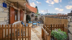 Un bâtiment en pierre avec une porte en bois, un espace extérieur couvert avec des coussins bleus, et une plante en pot.