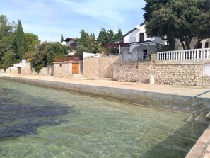 A swimming pool with clear water, a stone wall, and houses with balconies. - Amazing 2 Storey Holiday Home With 120m Roof Terrace, Directly on the Beach (Sveti Filip i Jakov)