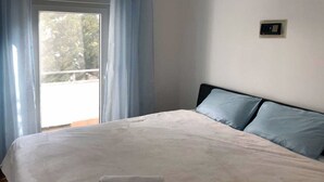 Ein ordentlich bezogenes Bett mit weißer Decke und zwei Kissen, ein Fenster mit blauen Vorhängen und eine wandmontierte Steuerung.