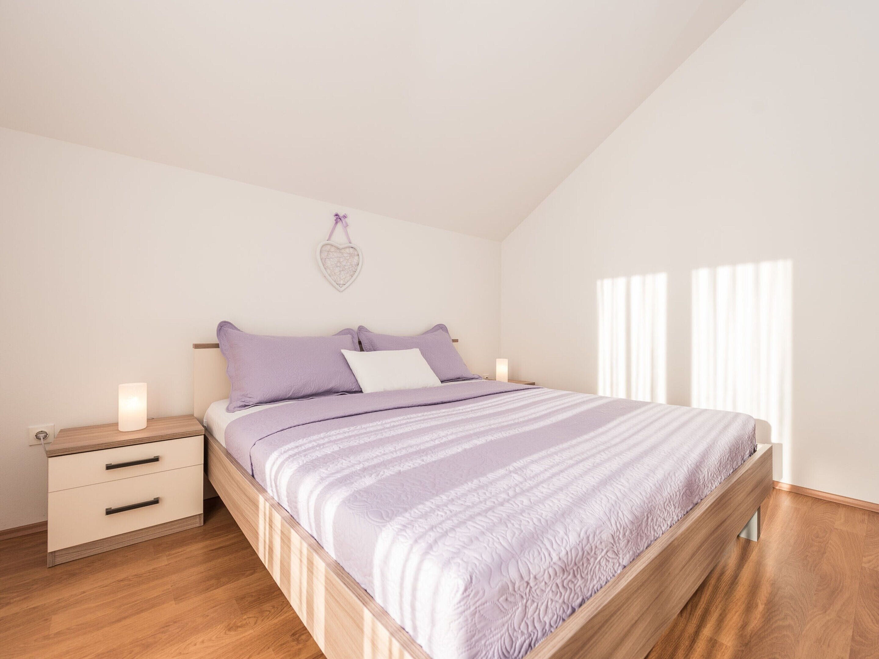 A bedroom with a bed, wooden nightstands, a drawer unit, and a heart-shaped wall decoration.