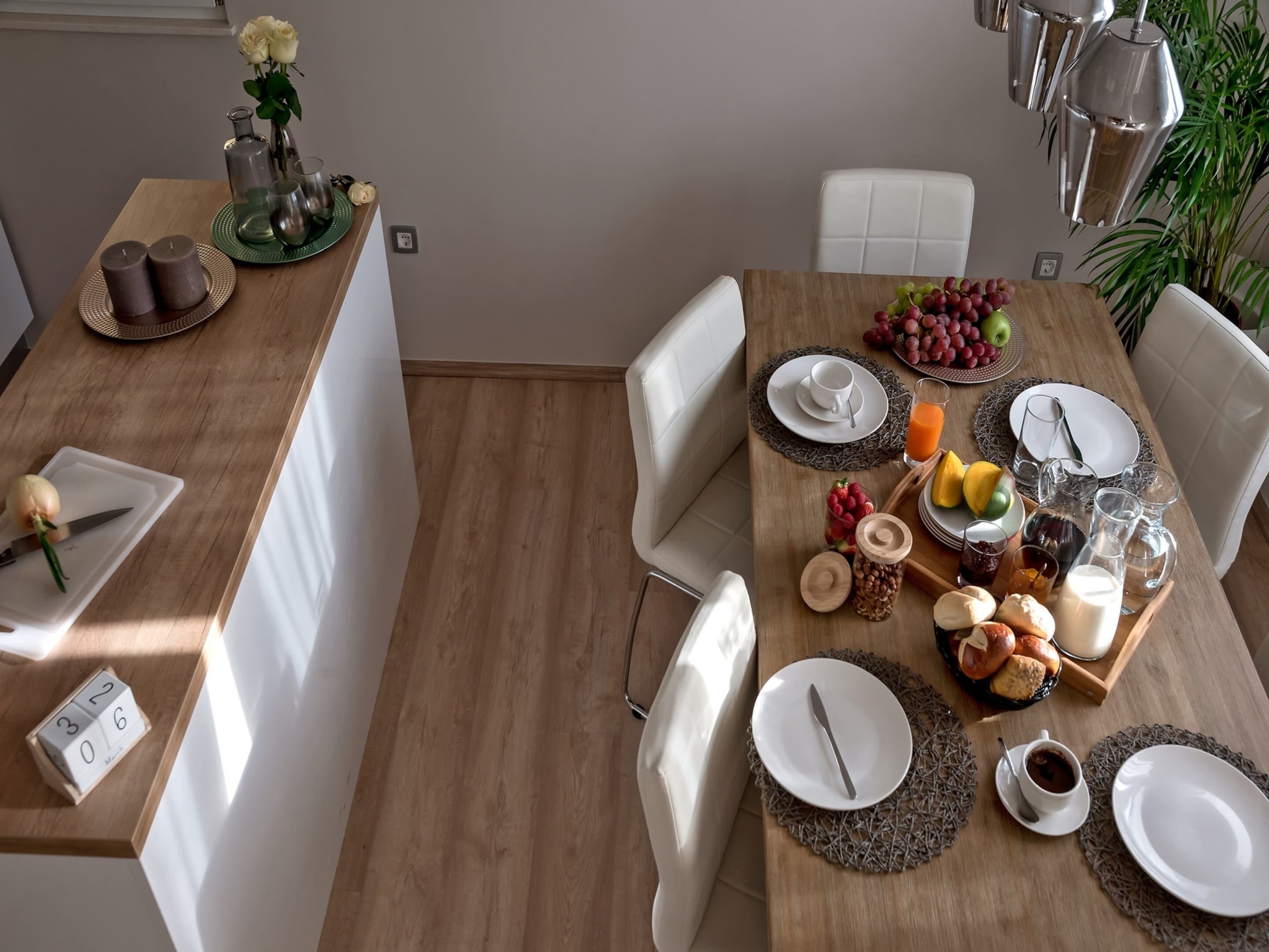 A dining area with a wooden table set for breakfast, including plates, cutlery, glasses, and a variety of fruits and pastries.