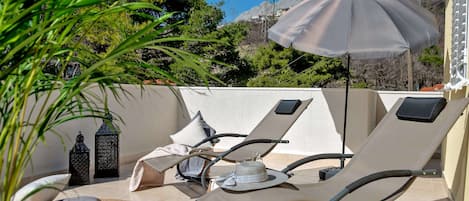 A sunlit outdoor seating area with lounge chairs and an umbrella, surrounded by greenery and a mountainous backdrop.