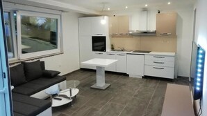 A modern kitchen with white cabinets, a built-in oven, and a stainless steel range hood. There is a dining table with a glass top and a black sofa in the living area. - Holiday Home in Crikvenica With Sea View (Crikvenica)