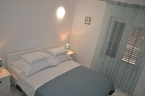A neatly made bed with pillows and a blue blanket, a bedside lamp, and a window with blinds. - Apartment in Molat With sea View (Zadar)