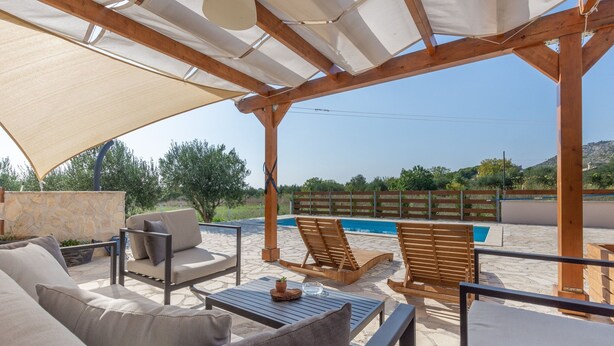 Un patio exterior con piscina, mobiliario de madera y una pérgola que proporciona sombra.
