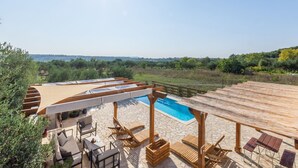 Zona de piscina con tumbonas de madera y una pérgola, rodeada de baldosas de piedra y vegetación.