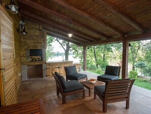 A covered patio area with wooden furniture, a stone fireplace, and a view of a lake. - Amazing Chalet with Private Garden, Hot Tub, Sauna, Great Location by the River (Lasinja)