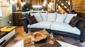 Un salon chaleureux avec un canapé incurvé, une table basse en bois et des verres à vin. On aperçoit, au fond, une cuisine dotée d’une cuisinière et d’un évier modernes.