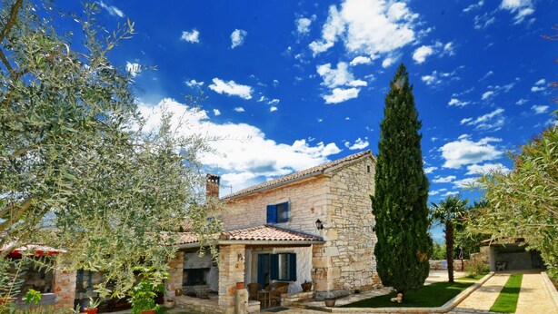 Une maison en pierre avec un toit en tuiles, une cheminée en pierre et un petit jardin agrémenté de plantes en pot et d’une pelouse.