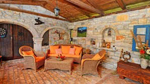 Une pièce rustique aux murs de pierre, au plafond en bois et au sol en briques. On y trouve un canapé avec des coussins orange, des chaises en osier et une commode en bois.