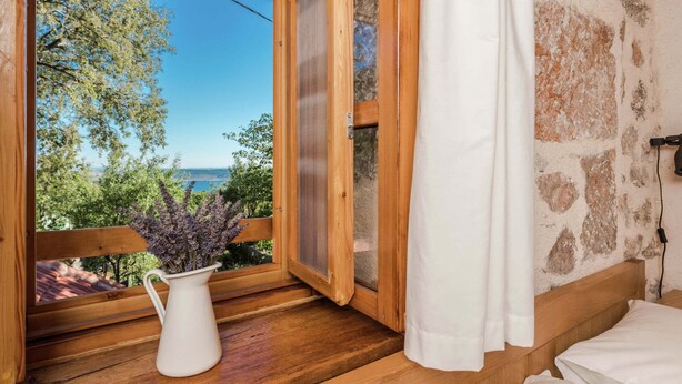 A room with a wooden floor, a white vase with flowers, a bed with white bedding, and a view of trees and a body of water through a large window. - Seaside Retreat in Marina di Grosseto (Starigrad)