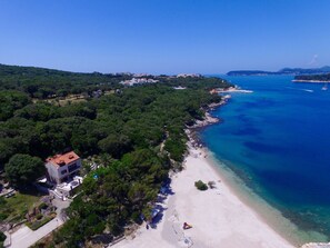 An aerial view of a coastal area with a beach, a building with a red roof, and a clear blue sea.