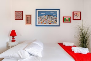 A hotel room with a bed, pillows, a red lamp, a plant, and several framed artworks on the wall.