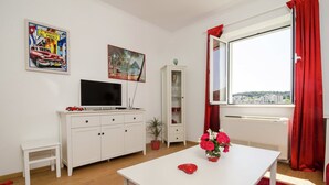 A room with a white table, red curtains, a TV on a white cabinet, a small white stool, a potted plant, and a window with a city view. - Dubrovnik Apartment With Private Terrace (Dubrovnik)