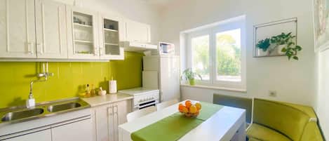 A compact kitchen with white cabinets, a green backsplash, a white countertop, and a window with a view of trees.