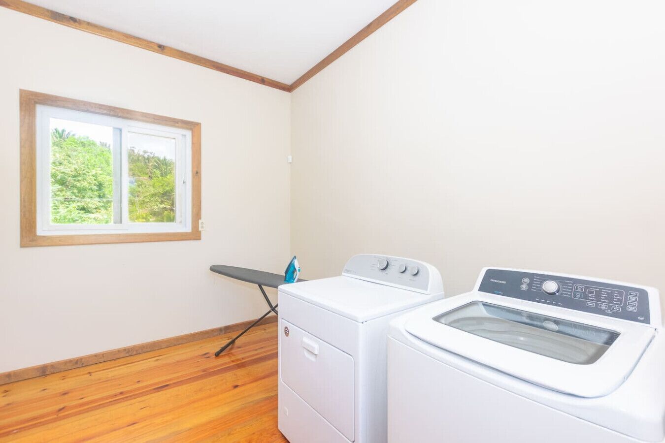 A laundry room with a washing machine, dryer, ironing board, and a window with a view of greenery.